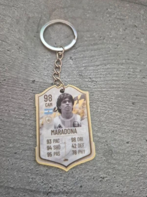 Nyckelring med Maradona-motiv - Säljer en unik nyckelring med Maradona-motiv inspirerad av FIFA Ultimate Team-kort. Nyckelringen är i plast och metall, med svartvitt porträtt och guldiga detaljer. Perfekt för dig som älskar fotboll och vill ha en cool accessoar till nycklarna.