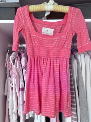 Rosa randig topp från Abercrombie & Fitch - Supersöt rosa topp från Abercrombie & Fitch med vita ränder, smockad midja och trekvartsärm. Toppens material känns mjukt och stretchigt, perfekt för en chill och trendig look. Snygg fyrkantig ringning och lätt utställd passform.