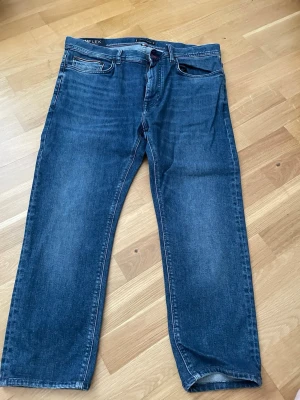 Mörkblå jeans från Tommy Hilfiger - Snygga mörkblå jeans från Tommy Hilfiger med TH Flex advanced stretch för extra komfort. Klassisk femficksmodell med raka ben och diskreta röda och vita detaljer på insidan. Perfekta för en avslappnad men stilren look. Storlek 36/32 .OBS! De har blivit förkortade med någon cm, fråga om exakt längd vid intresse så skulle säga snarare de är 36/30 eller 36/28
