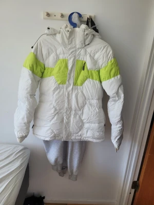 Vit dunjacka med limegröna detaljer - Säljer en vit dunjacka med limegröna detaljer över bröst och armar. Jackan har huva med dragsko, dragkedja framtill och två sidofickor. Perfekt för dig som vill ha en varm och snygg jacka till vintern. Materialet är klassiskt dun för maximal värme.