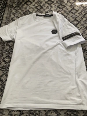 Vit Philipp Plein t-shirt med logga - Säljer en clean vit t-shirt från Philipp Plein med svart logomärke på bröstet och en snygg svart patch med guldfärgad text på ärmen. Klassisk passform och rund halsringning. Perfekt för dig som gillar exklusiva streetwear-plagg.