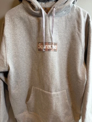 Grå Supreme x Burberry hoodie - Säljer en grå hoodie från Supreme x Burberry med klassiskt rutigt Supreme-tryck på bröstet. Hoodien har huva med dragsko och stor magficka. Tillverkad i mjuk bomullsblandning och har en avslappnad passform. Perfekt för dig som gillar streetwear och exklusiva samarbeten.