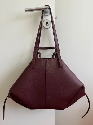 Vinröd shoppingväska i läder - Snygg shoppingväska i vinrött läder med unik geometrisk form och långa handtag. Väskan har minimalistisk design och diskreta sömmar, perfekt för dig som vill ha en stilren och rymlig accessoar. Passar till det mesta och rymmer mycket.