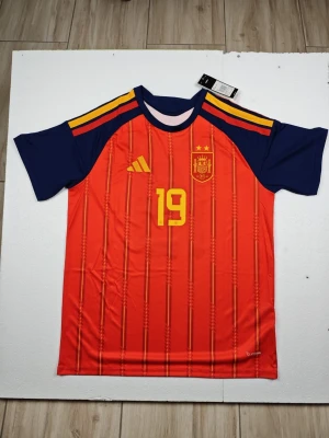 Spaniens matchtröja Lamine Yamal #19 - Röd och marinblå matchtröja från Adidas för Spaniens landslag med gula detaljer, nummer 19 och Lamine Yamal på ryggen. Tröjan har korta ärmar, rund hals och tre gula ränder på axlarna. Tillverkad i lätt och ventilerande funktionsmaterial.