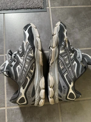 Svarta och grå Asics sneakers - Säljer ett par svarta och grå sneakers från Asics med klassisk mesh och syntetmaterial. Skorna har en sportig design med tydliga Asics-loggor på sidan och en tjock, dämpad sula. Perfekta för dig som gillar streetwear och bekväma skor.