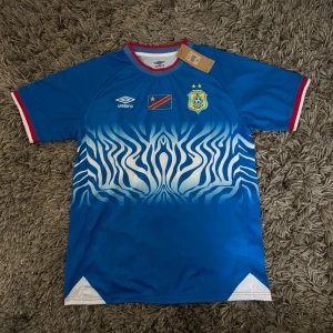 Blå Umbro fotbollströja DR Kongo - Säljer en blå fotbollströja från Umbro med DR Kongos flagga och förbundsmärke på bröstet. Tröjan har ett vitt zebramönster framtill, röda och vita detaljer vid krage och ärmslut samt två stjärnor ovanför klubbmärket. Perfekt för match eller träning.