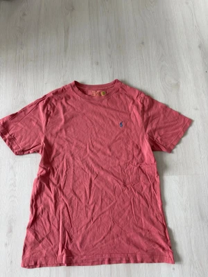 Rosa t-shirt från Polo Ralph Lauren - Snygg rosa t-shirt från Polo Ralph Lauren med klassisk passform och rund hals. T-shirten har en liten blå broderad logga på bröstet och är tillverkad i mjuk bomull. Perfekt för en avslappnad och stilren look.