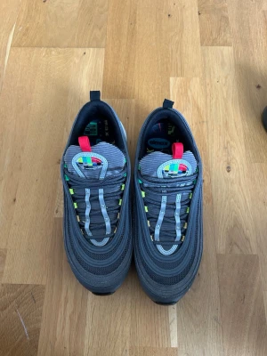 Nike Air Max 97 grå sneakers - Nike Air Max 97 sneakers i grått med reflekterande detaljer, färgglada accenter på plösen och innersulan med Air Max-loggor. Skorna har vågformade linjer, mesh och syntetmaterial samt synlig Air Max-dämpning i sulan. Kommer utan sin original box!