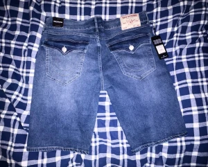 Blå jeansshorts från True Religion - Säljer ett par klassiska blå jeansshorts från True Religion med ikoniska bakfickor och kontrastsömmar. Shortsen har normal passform, knappgylf och fem fickor. Perfekta för sommaren och riktigt snygga till en enkel t-shirt.