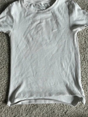 Vit ribbad t-shirt med kort ärm - En enkel och stilren vit t-shirt med ribbad struktur och korta ärmar. Perfekt basplagg till alla outfits och passar året runt. T-shirten har en rund halsringning och är gjord i mjukt material för skön komfort. (Liten fläck upptill)