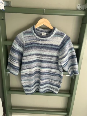 Blåmelerad stickad tröja Stories XS - Säljer en blåmelerad stickad tröja från Stories i storlek XS (32-34). Tröjan har rund halsringning, korta ärmar och ett randigt mönster i olika blå och grå toner. Perfekt för dig som gillar en avslappnad och mysig stil.