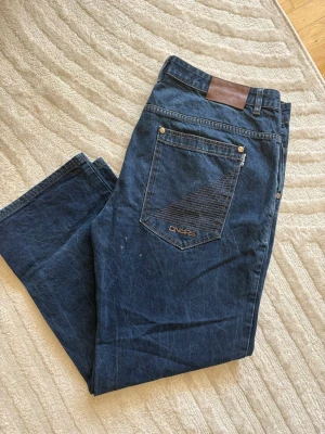Baggy jeans från DNGRS - Säljer ett par mörkblå jeans från DNGRS med raka ben/baggy fit och klassisk femficksmodell. Jeansen har snygga kontrastsömmar och en diskret logga på bakfickan & framsidan. Defekter - På första bilden syns svagare fläckar, de är obehandlade & går säkert bort med fläckmedel. Storlek - 44/32 