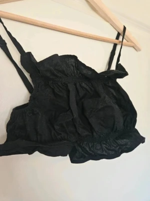 Svart linnetopp med volangdetaljer - Trendig svart croppad linnetopp med tunna justerbara axelband och volangdetaljer. Toppen har en luftig passform och elastiska partier för extra komfort. Perfekt att styla med höga jeans eller kjol. E4