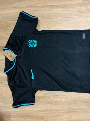 Svart Brasilien fotbollströja Nike - Snygg svart Brasilien fotbollströja från Nike med turkosa detaljer och Dri-FIT-material. Tröjan har rund hals med turkos kant och matchande detaljer på ärmsluten. Perfekt för träning eller match!