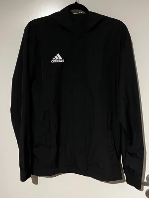 Svart vindjacka från Adidas - Snygg svart vindjacka från Adidas med vit logga på bröstet. Jackan har dragkedja framtill, två sidofickor och en enkel, sportig design. Perfekt för blåsiga dagar och passar till det mesta i garderoben.