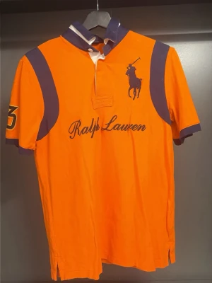 Orange Ralph Lauren t-shirt - Snygg orange t-shirt från Ralph Lauren med mörkblå detaljer på axlar, ärmslut och krage. Stort logotryck och text på bröstet samt siffra på ena ärmen. Klassisk pikékrage med knappar och sportig vibe.