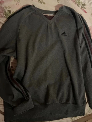 Grå v-ringad tröja från Adidas - Mysig grå tröja från Adidas med v-ringning och diskret logga på bröstet. Tröjan har långa ärmar med mörka ränder längs sidorna och ribbade muddar vid ärmslut och nederkant. Perfekt för en sportig och avslappnad stil.
