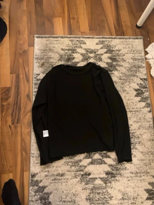 Svart långärmad tröja från 157 - En enkel svart långärmad tröja från 157 med rund halsringning. Tröjan är i ett mjukt och stretchigt material som passar perfekt till alla typer av outfits. Stilren och lätt att matcha med andra plagg.