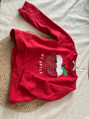 Röd sweatshirt med paljettäpple - Röd sweatshirt med långa ärmar och rund hals. Framtill finns ett stort paljettäpple i rött, vitt och grönt samt texten 'MY APPLE'. Tröjan är gjord i mjukt bomullstyg och har ribbade muddar vid ärmslut och nederkant. Perfekt för dig som gillar färgglada detaljer. Storlek 98/104