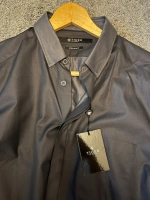 Svart skjorta från Tiger of Sweden - Stilren mörkblå skjorta från Tiger of Sweden med klassisk krage och knappar framtill. Skjortan har lång ärm och är tillverkad i ett mjukt bomullstyg som ger en clean och modern look. Perfekt för dig som gillar minimalistisk stil. 186