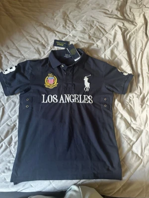 Mörkblå piké Los Angeles Polo Ralph Lauren - Mörkblå pikétröja från Polo Ralph Lauren med vit text 'LOS ANGELES' och broderad logga på bröstet. Nummer 3 på ryggen och ärmen, samt detaljer i vitt och gult. Klassisk krage och korta ärmar.