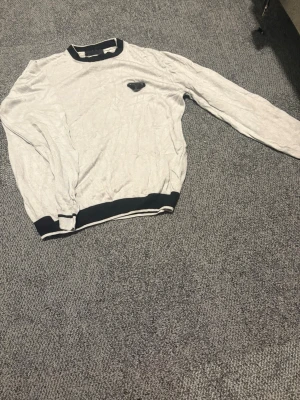 Ljusgrå tröja med svarta detaljer - Ljusgrå långärmad tröja med svarta ribbade muddar vid hals, ärmslut och nederkant. Tröjan har en liten svart logotyp på bröstet och är tillverkad i ett tunt, mjukt material. Enkel och stilren design som passar till det mesta.