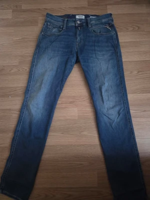 Blå slim fit jeans från Replay - Säljer ett par blå slim fit jeans från Replay med klassisk femficksdesign och snygga slitningar. Jeansen har normal midja och smala ben, perfekta för en avslappnad men ändå stilren look. Materialet är mjukt denim som sitter skönt hela dagen.