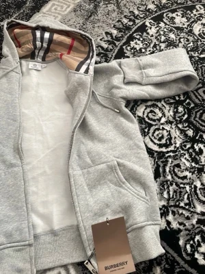 Grå hoodie med dragkedja från Burberry - Snygg grå hoodie från Burberry med dragkedja och klassiskt rutigt foder i huvan. Tröjan har fickor framtill och ribbade muddar vid ärmslut och nederkant. Perfekt för en avslappnad och trendig look. Passar S eller Liten M. Säljer då jag fick den som present och är alldeles för liten för mig.