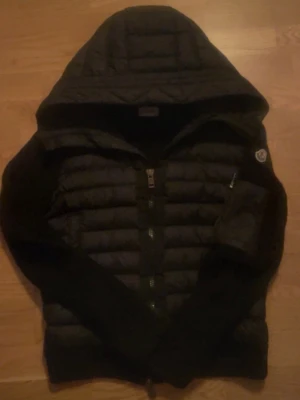 Svart Moncler pufferjacka med huva - Svart pufferjacka från Moncler med stor huva, quiltad kropp och ribbstickade ärmar. Jackan har dragkedja framtill och Moncler-logga på ärmen. Materialet är en mix av vadderad nylon och stickat tyg för extra komfort och värme.