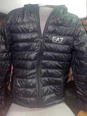 Svart dunjacka från EA7 Emporio Armani - Snygg och stilren EA7 Emporio Armani pufferjacka i svart. Jackan har EA7-logga på bröstet, quiltad/puffad design och huva. Perfekt för både vardag och en clean streetwear-look. Märke: EA7 Emporio Armani Storlek: M Färg: Svart Modell: Puffer/dunjacka med huva Detaljer: EA7-logga på bröstet och EA7-detalj på dragkedjan Skick:se bilder för egen bedömning 📦 Skickas snabbt och packas ordentligt. 💬 Hör gärna av dig vid frågor