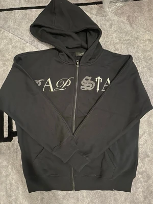 Svart hoodie med dragkedja och tryck - Säljer en svart hoodie med dragkedja och huva. På bröstet finns ett tryck i olika typsnitt som stavar 'TRAPSTAR'. Insidan har även ett tryck med bokstäverna 'TS'. Hoodien har ribbade muddar och fickor framtill. Materialet känns mjukt och bekvämt.