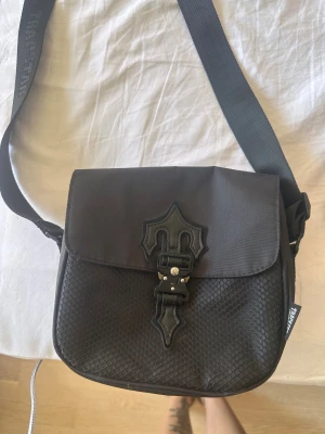 Svart axelväska från Trapstar - Snygg svart axelväska från Trapstar med justerbar rem och coolt spänne framtill. Väskan har en unik struktur med hexagonmönster på framsidan och detaljer i konstläder. Perfekt för dig som vill ha en streetwear-inspirerad look. 
