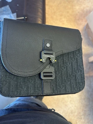 Svart axelremsväska  - Snygg svart axelremsväska med präglat monogramtyg och detaljer i läder. Väskan har ett coolt spänne framtill med logotyp och justerbar rem. Perfekt för dig som vill ha en stilren och exklusiv look.