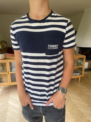 Tommy Hilfiger randig T-shirt herr - Tommy Jeans T-shirt i stilren marinblå/vit randig design. Bröstficka med Tommy Jeans-logga och klassisk rund hals. Snygg och lättmatchad modell, perfekt till jeans eller shorts. Storlek S. Fint skick.  #tommyjeans #tommyhilfiger #tshirt #randig #striped #marinblå #streetwear #preppy #casual #vintage
