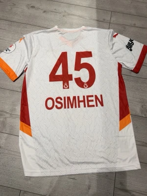 Galatasaray bortatröja Puma - Snygg Galatasaray bortatröja från Puma i vitt med röda och orange detaljer. Tröjan har nummer 45 och namnet Osimhen på ryggen, samt klubbmärke och sponsortryck på framsidan. Tillverkad i lätt och ventilerande material, perfekt för fotboll eller träning.