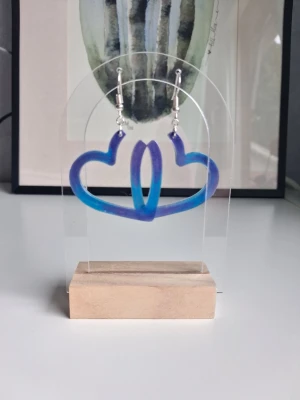 Blå-lila hjärtformade örhängen resin/epoxy  - Unika örhängen gjorda i resin/epoxy i blå och lila skimmer/metallisk färg med öppen hjärtform. De har silvriga krokar som är nickelsäkraoch en cool, modern design som verkligen sticker ut. Perfekta för dig som vill ha något färgglatt och lekfullt i din stil. Längd ca 4.5cm långa