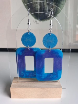 Blå-lila statement örhängen i resin/epoxy - Handgjorda örhängen i resin/epoxy med stora rektangulära former i blått och lila marmorerat skimmer mönster. Örhängena har en rund blå platta upptill och hängs i silverfärgade krokar som är nickelsäkra. Perfekta för dig som vill sticka ut med färg och cool design. Längd ca 5.3cm långa.