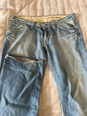 Ljusblå jeans från Replay - Säljer ett par ljusblå jeans från Replay med klassisk femficksmodell och slitningar på ena benet. Jeansen har normal midja, raka ben och detaljerade sömmar. Perfekta för dig som gillar en avslappnad och trendig stil.