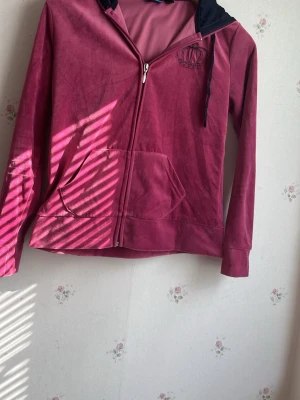 Mörkrosa hoodie med kronbrodyr - Säljer en vintage mörkrosa hoodie med dragkedja och stor ficka framtill. Huvan har mörkblå insida och snören. På bröstet finns en broderad krona i svart. Materialet ser ut att vara mjuk velour, perfekt för en chill och avslappnad stil.