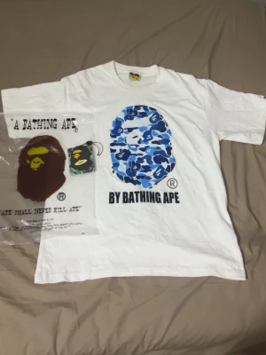 Vit BAPE t-shirt med blått camotryck - Säljer en vit t-shirt från A Bathing Ape med det klassiska BAPE-huvudet i blått camomönster på bröstet och texten 'BY BATHING APE' under. T-shirten har rund hals och korta ärmar. Perfekt för dig som gillar streetwear och vill sticka ut.