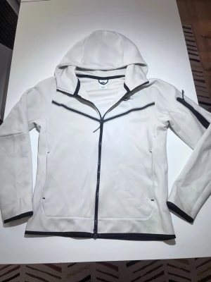 Vit hoodie med dragkedja från Nike - Snygg vit hoodie från Nike med hel dragkedja framtill och svarta detaljer längs dragkedjor och muddar. Hoodien har en huva och fickor på sidorna samt en extra ficka på ärmen. Tillverkad i mjukt material, perfekt för en sportig och avslappnad stil.