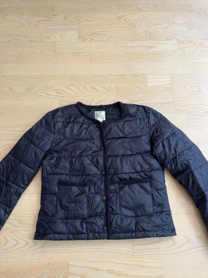 Mörkblå kraglöspufferjacka - Snygg mörkblå pufferjacka med rund halsringning och utan krage. Jackan har två stora framfickor och stängs med knappar framtill. Quiltad design och lätt vadderad för extra värme. Perfekt för kalla dagar och enkel att matcha med olika outfits.