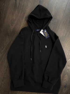 Svart hoodie från Polo Ralph Lauren - Snygg svart hoodie från Polo Ralph Lauren med klassisk huva och dragsko. Ikoniska vita polospelaren broderad på bröstet. Mjuk bomullskänsla och känguruficka framtill. Perfekt för en clean och stilren look.