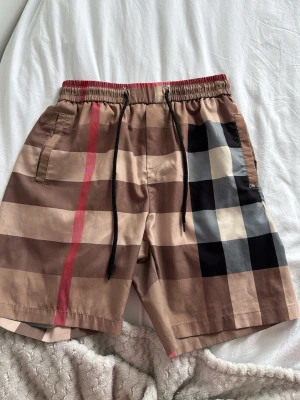 Rutiga shorts med snörning - Snygga shorts med klassiskt rutigt mönster i beige, brunt, svart och rött. De har elastisk midja med svart snörning och sidofickor. Perfekta för sommaren och ger en trendig vibe till din outfit.