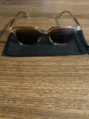 Raybandliknande solglasögon  - Snygga solglasögon med transparent, fyrkantig båge och mörka glas. Bågarna har en lätt beige ton och glasen är mörkbruna. Perfekta för soliga dagar och ger en cool vibe till din outfit. Kommer med en svart tygpåse för förvaring. Ska vara uv skydd men har ej testat för uv skydd 