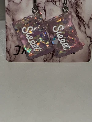 Lila Sweets’ Örhängen  - Unika örhängen i form av Sweets’ med lila bas, fyllda med holografiskt glitter och texten 'Sweet' i vitt. De har en lekfull och färgglad design som verkligen sticker ut . Örhängens krokarna är i rostfritt stål 