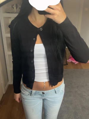 Svart cropped kofta med knappar - Snygg svart cropped kofta med rund halsringning och knäppning framtill. Långärmad och i tunt, mjukt material som sitter tajt. Perfekt att styla över ett linne eller t-shirt för en trendig look.