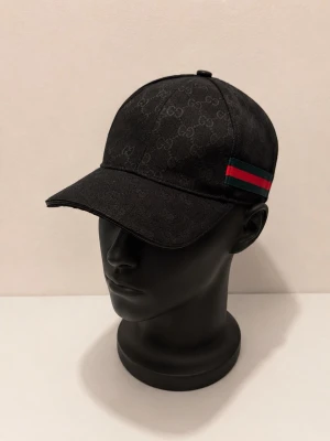 Svart randig keps med GG mönster  - Snygg svart keps fmed klassiskt GG-monogram över hela kepsen och en ikonisk grön-röd rand på sidan. Kepsen har böjd skärm och justerbar rem bak. Tillverkad i ett slitstarkt tyg med exklusiv känsla och detaljer med GG-logga på insidan.