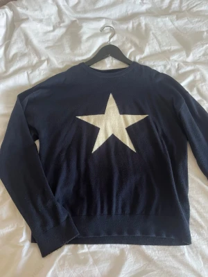 Mörkblå stickad tröja med stjärna - Snygg mörkblå stickad tröja med en stor vit stjärna på bröstet. Tröjan har rund halsringning och långa ärmar. Perfekt för dig som gillar enkel men cool design. Materialet är mjukt och bekvämt i viskos, polyamid & polyester.