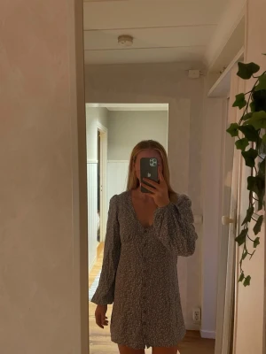 Blommig kortklänning med v-ringning - Söt kortklänning med blommigt mönster i blått, vitt och gult. Klänningen har v-ringning, långa ärmar och en figurnära passform. Perfekt för sommarens alla tillfällen och ger en fräsch och trendig look.